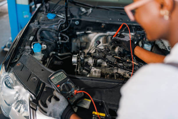 A young African American female mechanic is standing next to a car with a popped hood and repairing the car battery.