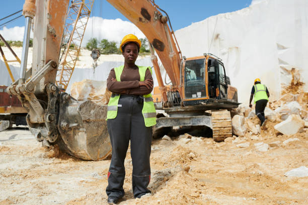 Young serious African American woman in workwear standing on construction site against caterpillar truck and her colleague