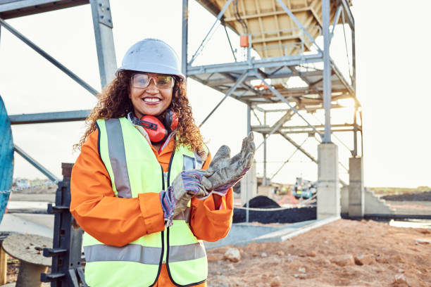 A young African woman mine worker wearing protective wear is looking at the camera while putting on a pair of gloves with coal mine equipment in the background